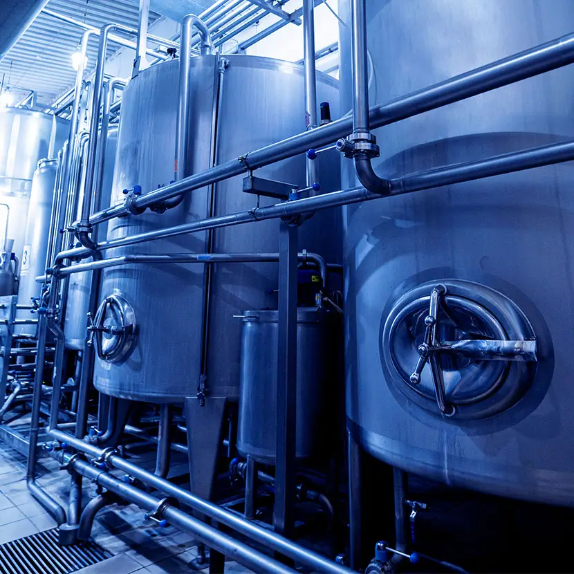 Industrial stainless steel tanks and pipes inside a modern beverage or food processing plant with blue-toned lighting.